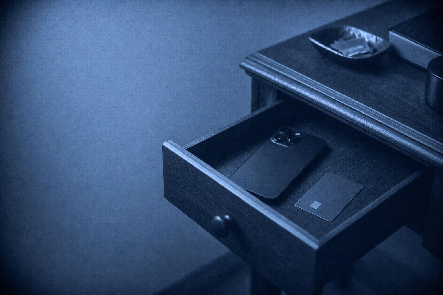 A desk drawer pulled half open with a phone lying face down inside it and a bank card beside it.